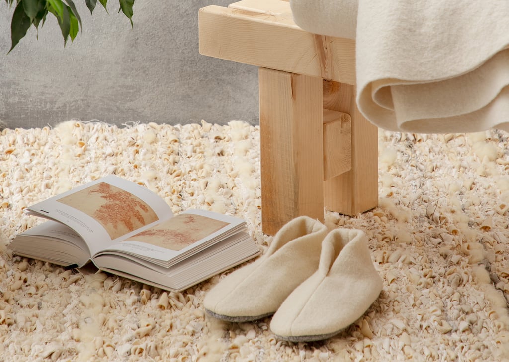 Cream slippers and an open book on a textured rug beside a wooden bed draped with a blanket.