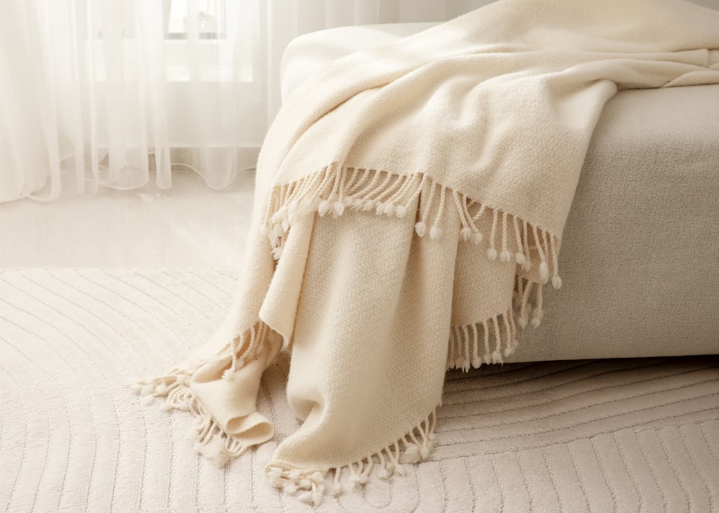 Cream blanket with knotted fringe draped over a light upholstered seat beside a sunlit window.