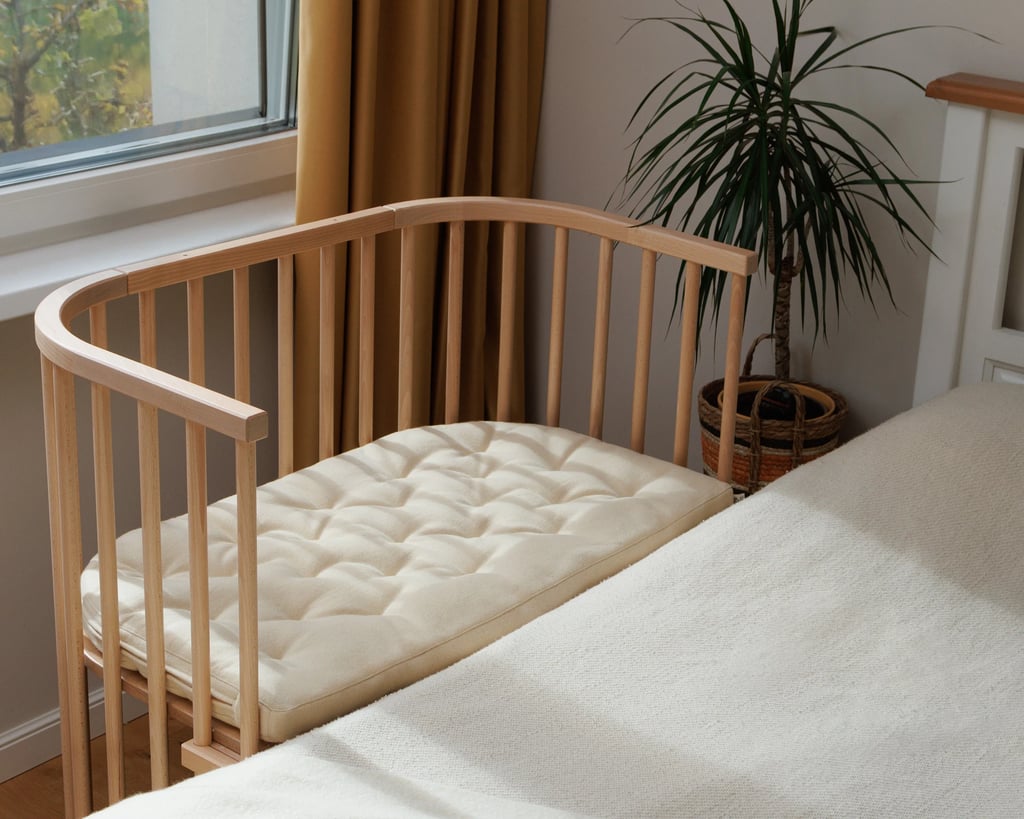 babybay-vugge i uld ved siden af en seng i et soveværelse med en plante og et vindue med brunt gardin i baggrunden