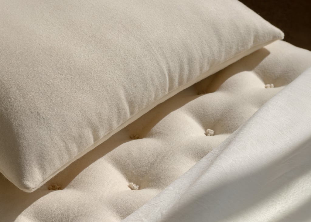 Close-up of a tufted cream mattress with a matching pillow and a white sheet in sunlight.
