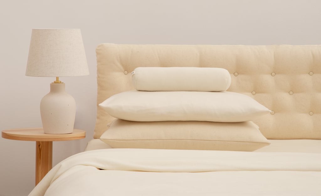 Two cream pillows and a cylindrical bolster on a bed beside a lamp on a wooden bedside table.