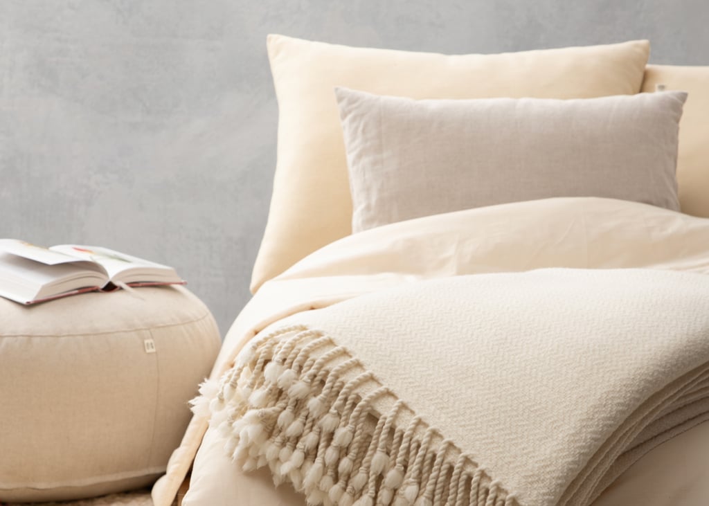 Bed layered with cream bedding, neutral pillows and a fringed blanket, beside an open book on a round pouf.