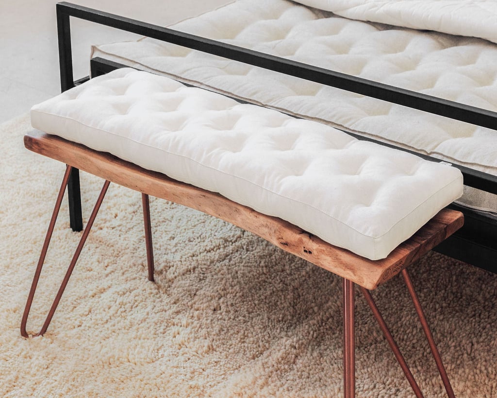 White tufted cushion on a wooden bench with dark metal legs beside a bed.