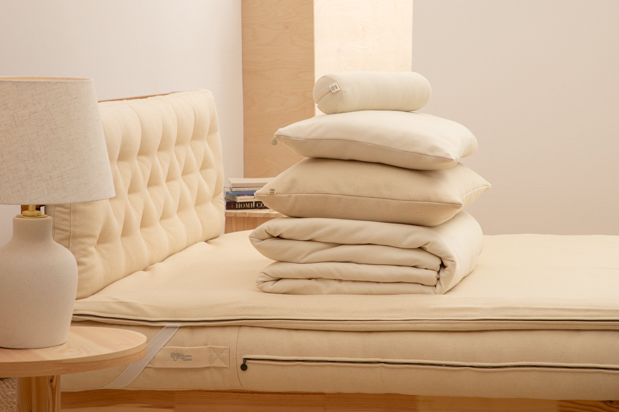 Cream mattress with stacked pillows, folded bedding and a bolster beside a tufted headboard.